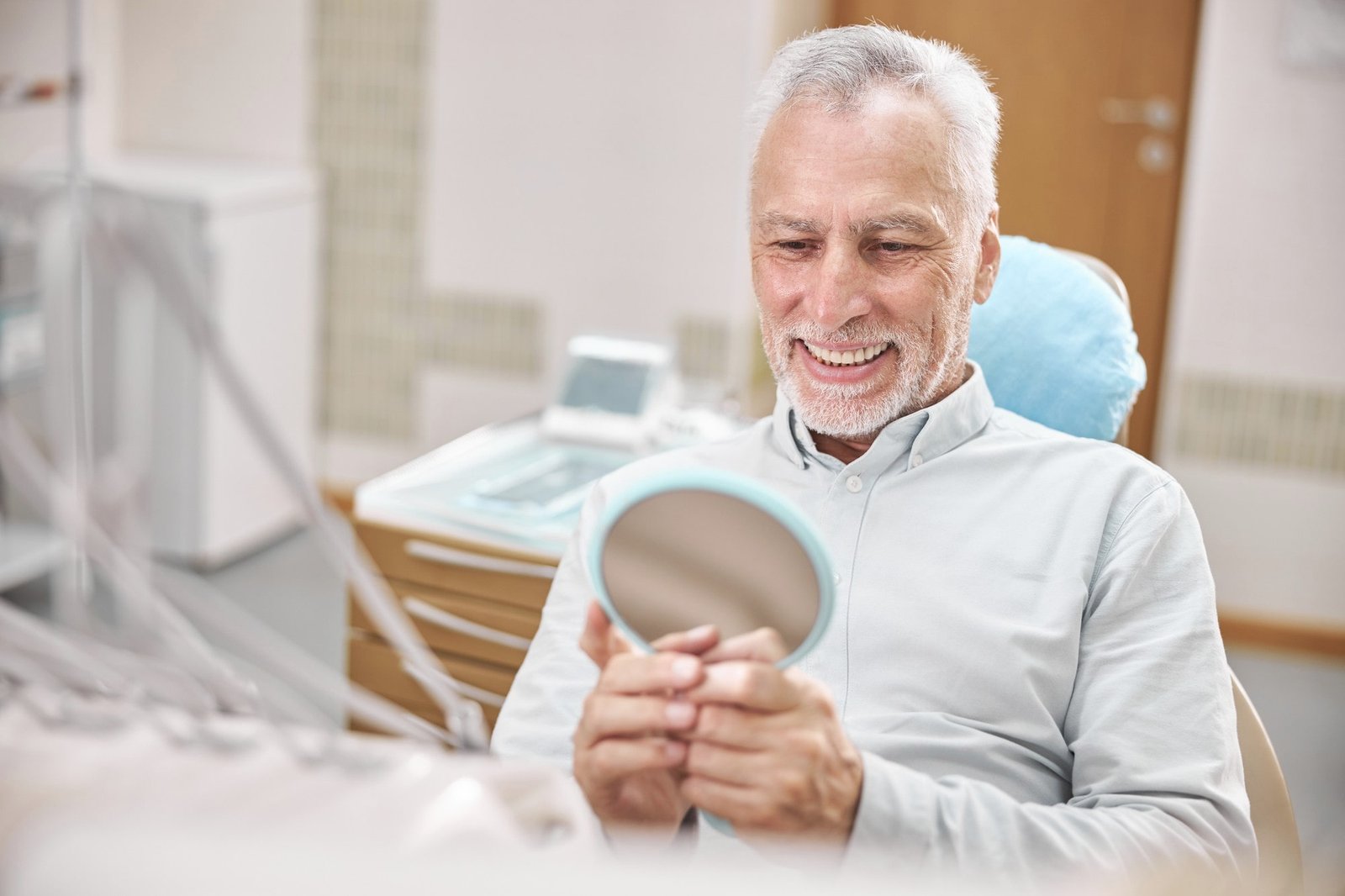 elderly-patient-dental-clinic-smiling-looking-mirror-while-sitting-dental-chair-min A close-up image of a person's smile with veneers showcasing perfectly aligned, white teeth and a natural-looking appearance.